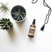 Image of Namaste Ninja Aromatherapy Mist on a white table with succulents. Brown fogged glass with kraft paper label. Available for purchase on The Conservationist.  